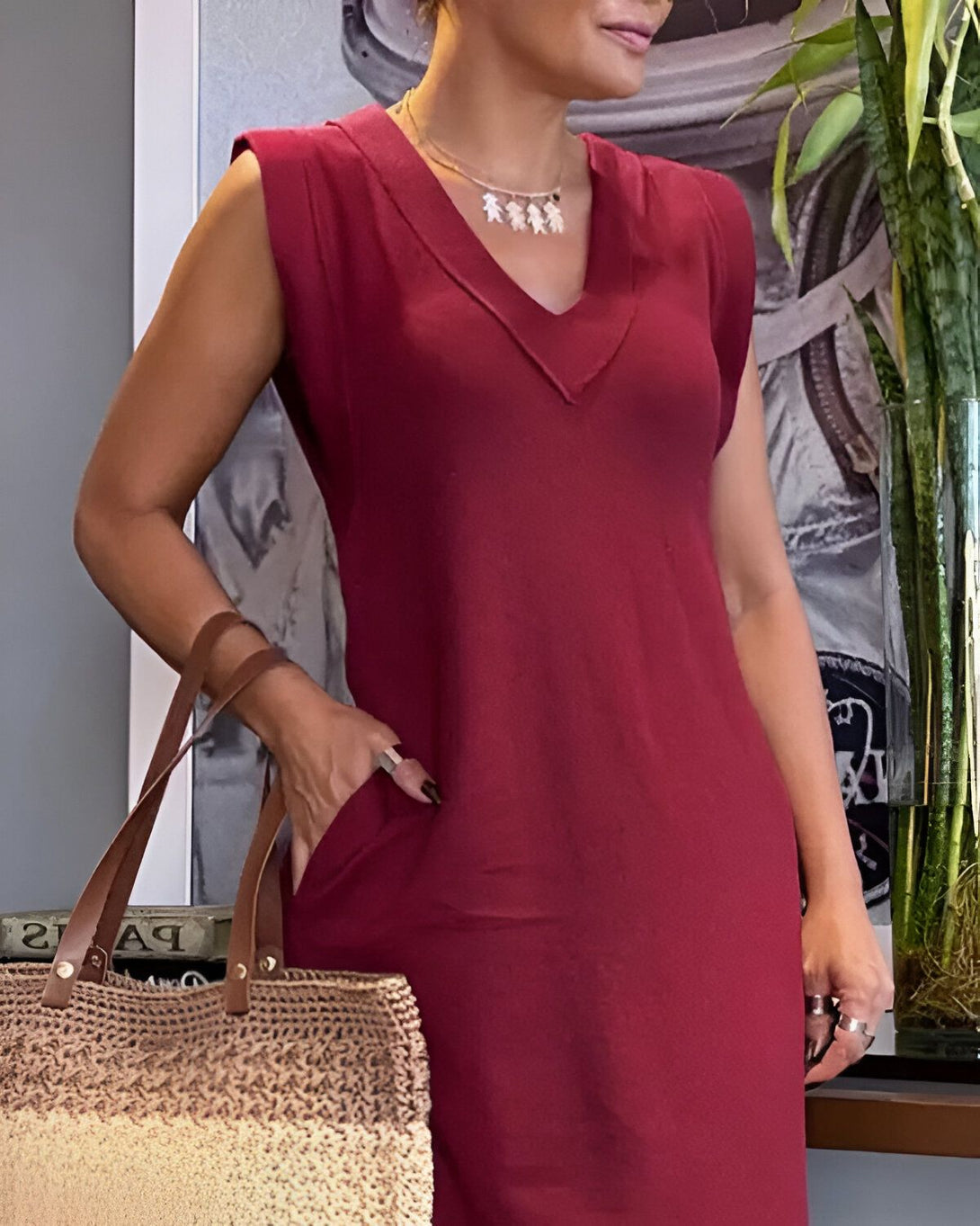 Une femme porte une robe rouge sans manches et un collier avec trois pendentifs. Elle a un sac à main en osier à la main droite et l'autre main dans la poche de sa robe.