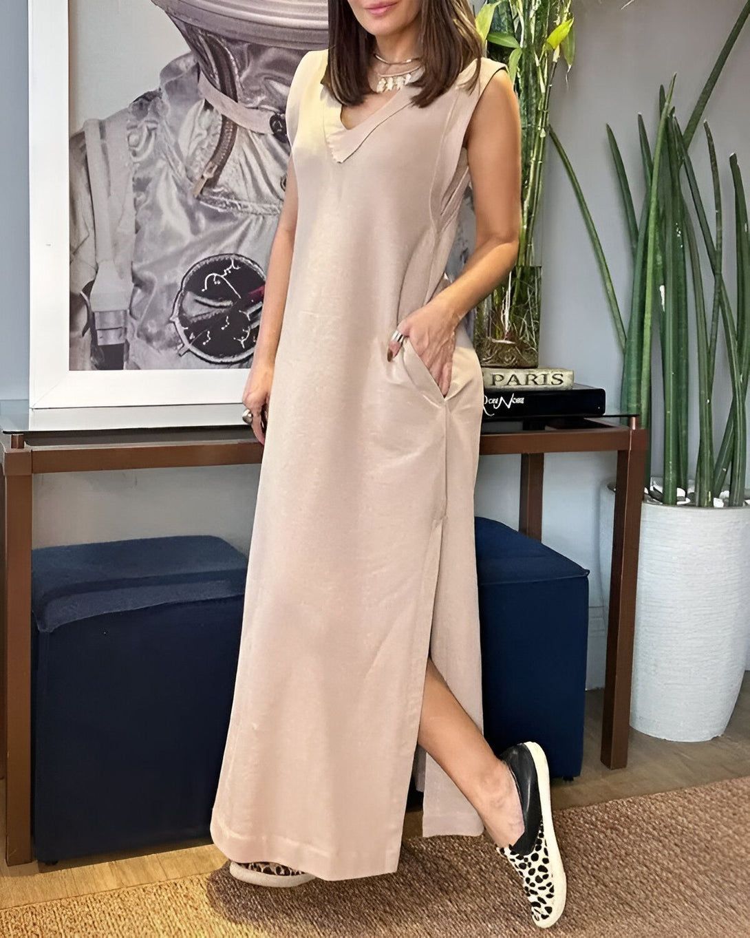 Une femme porte une robe longue beige sans manches avec des fentes latérales. Elle a des chaussures noires à motifs léopard et un collier. Elle se tient debout dans un salon, avec une main dans la poche et l'autre tenant un livre.