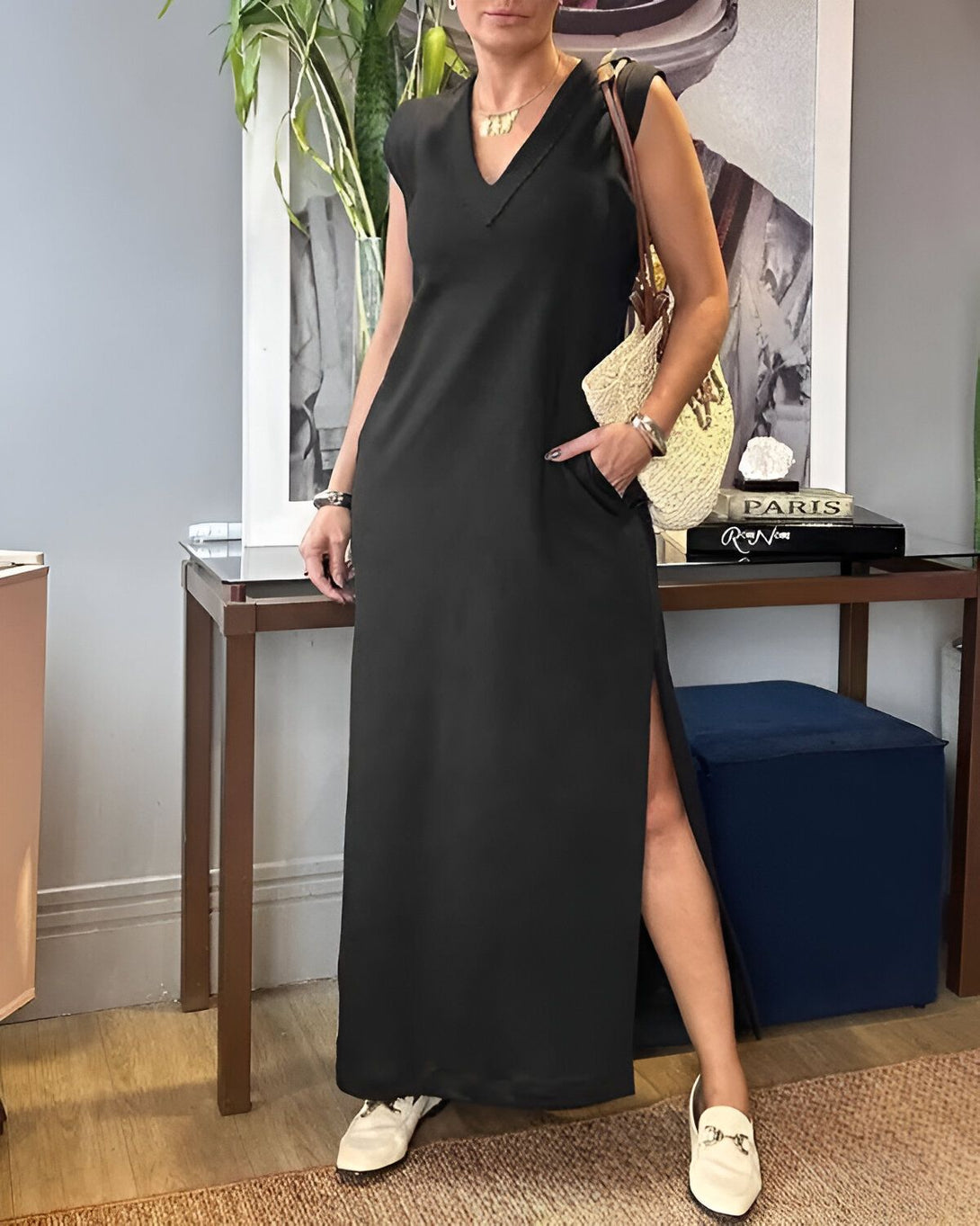 Une femme porte une longue robe noire sans manches avec une fente sur le côté. Elle a un sac à main en osier à la main et des chaussures blanches. Elle se tient debout dans un salon, il y a une plante verte et un tableau sur le mur.