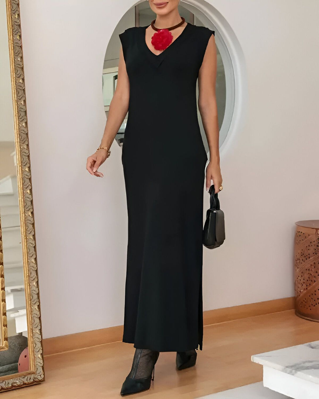 Une femme porte une robe noire longue et moulante, avec un décolleté en V. Elle porte également un collier rouge et un sac à main noir.