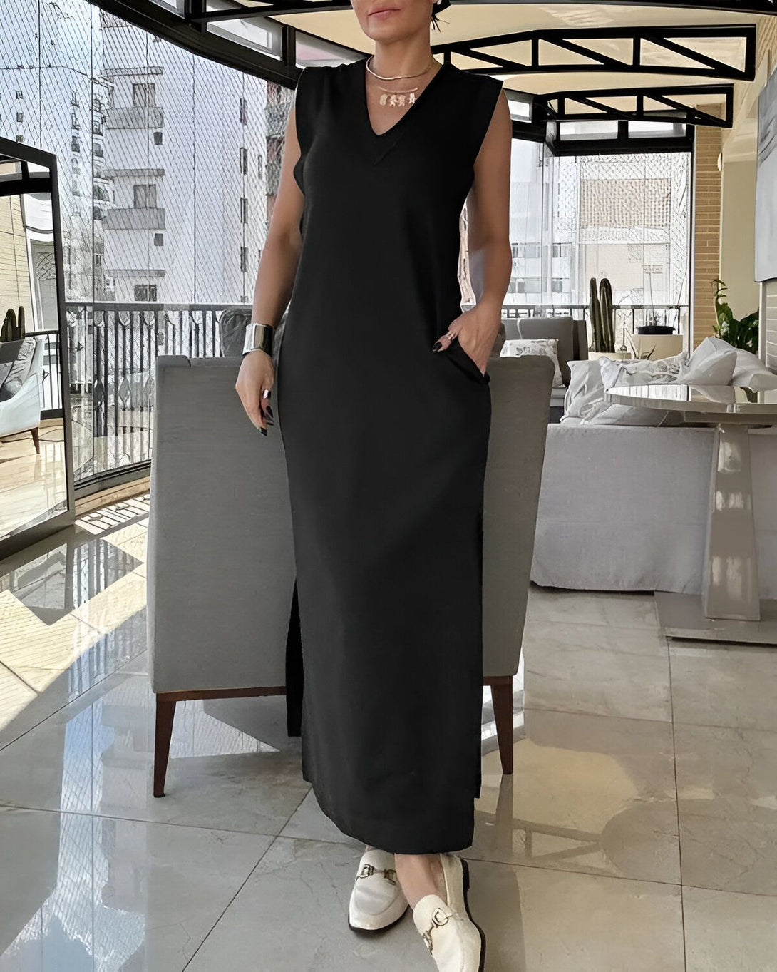 Une femme porte une longue robe noire sans manches avec des fentes sur les côtés. Elle a un collier et un bracelet en argent. Elle est debout dans un salon avec des murs en verre et un sol en marbre.