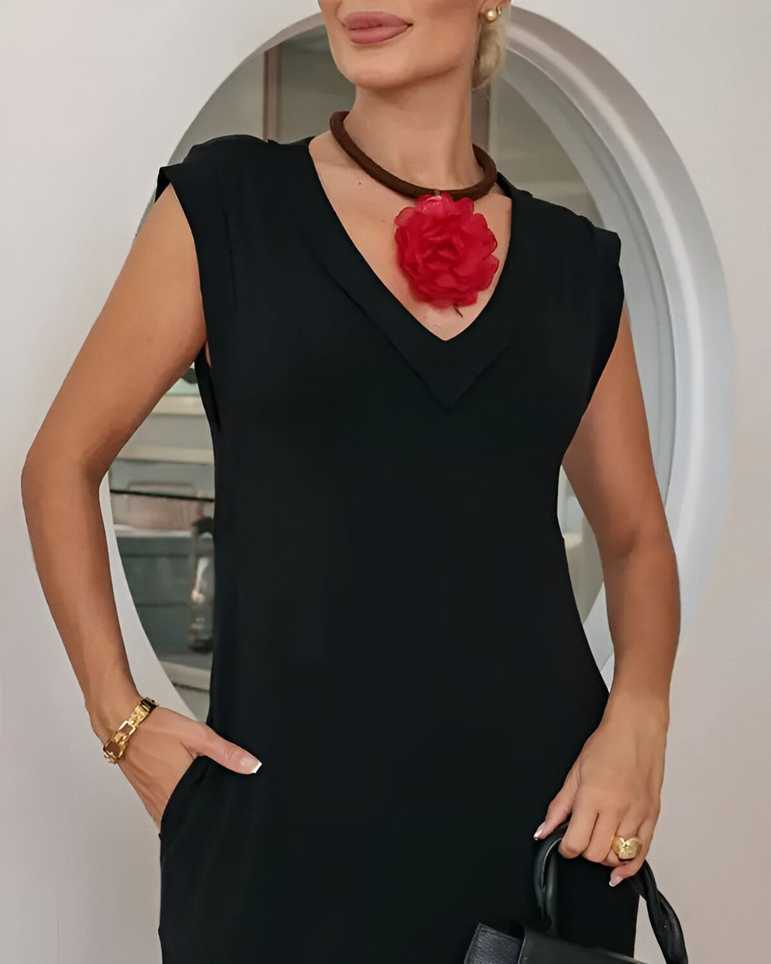 Une femme porte une robe noire sans manches avec un collier de fleur rouge en résine et un bracelet doré au poignet droit. Elle a une main dans la poche et tient un sac à main noir dans l'autre main.