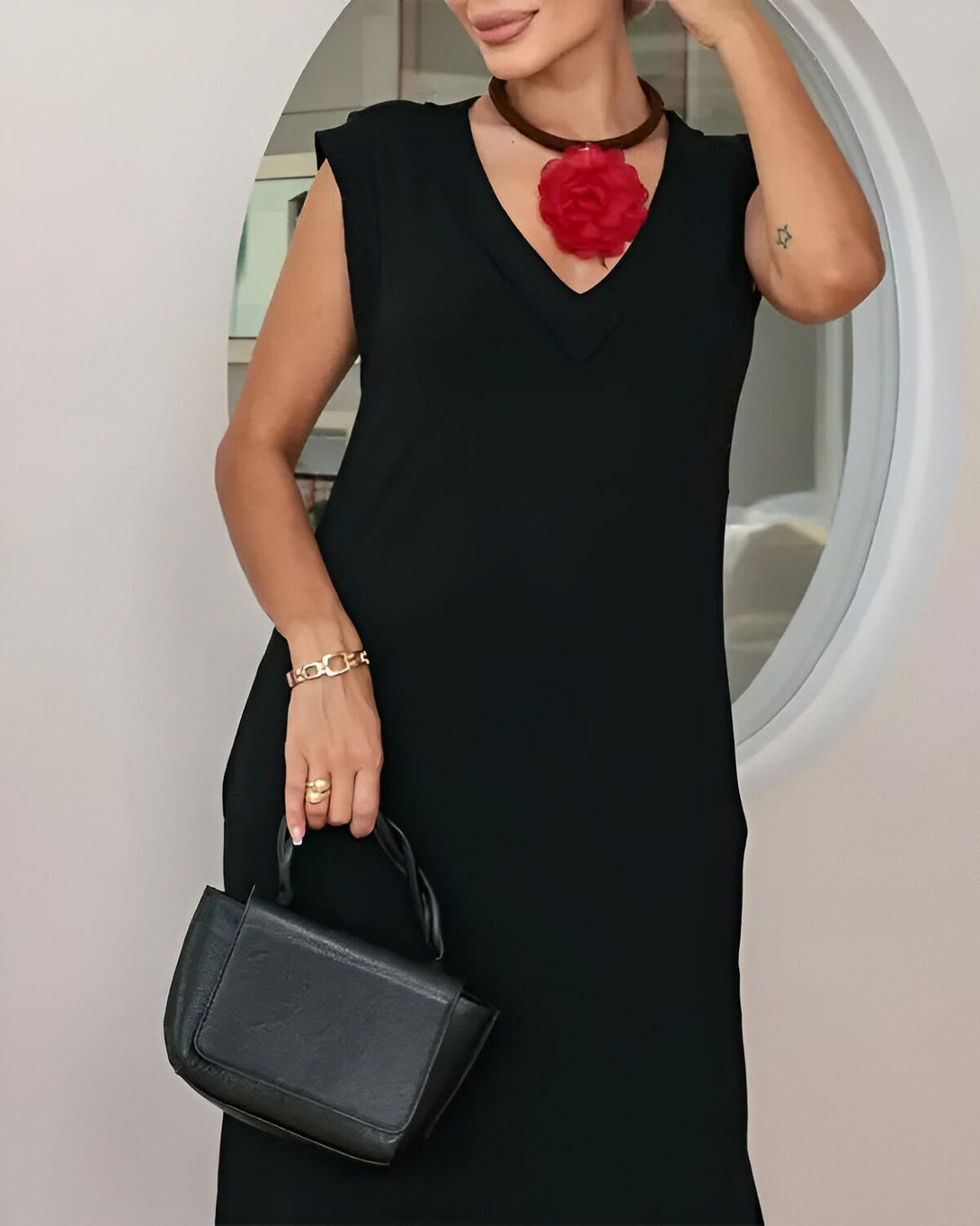 Une femme porte une robe noire sans manches et un collier de fleurs rouges. Elle tient un sac à main noir à la main.