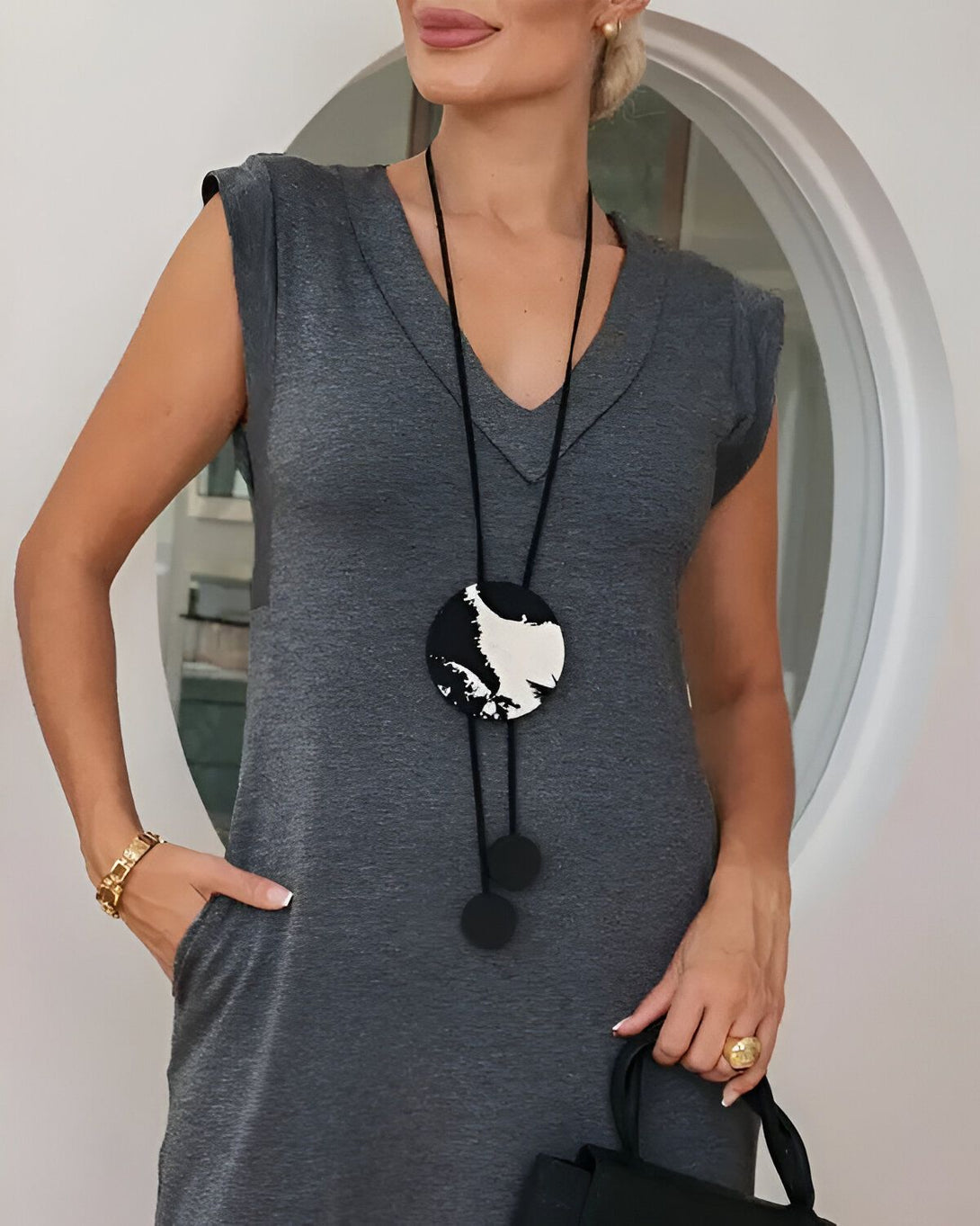 Une femme porte une robe en jersey de coton gris chiné à encolure V et manches chauve-souris. Elle porte également un collier pendentif rond en bois peint en noir et blanc et des boucles d'oreilles assorties. Elle a une montre en or au poignet gauche et une bague en or à l'annulaire droit. Elle tient un sac à main noir à la main droite.
