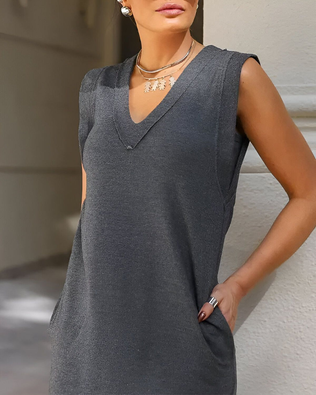 Une femme porte une robe droite gris anthracite sans manches, avec un décolleté en V. Elle porte également un collier argenté et des boucles d'oreilles en forme de feuilles.