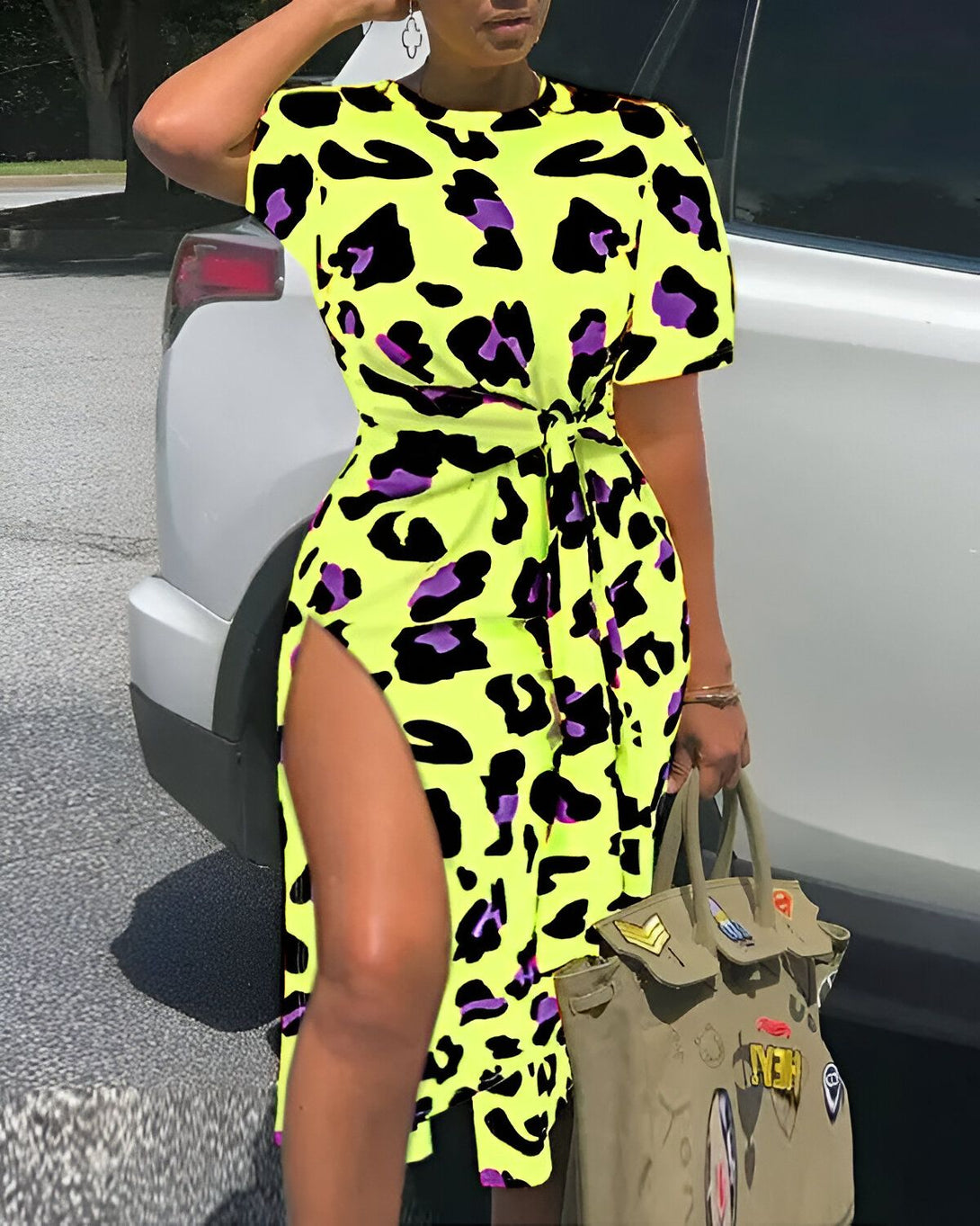 Une femme porte une robe jaune et noire à imprimé léopard. La robe est courte et fendue sur le côté. Elle porte également des sandales noires et un sac à main vert.