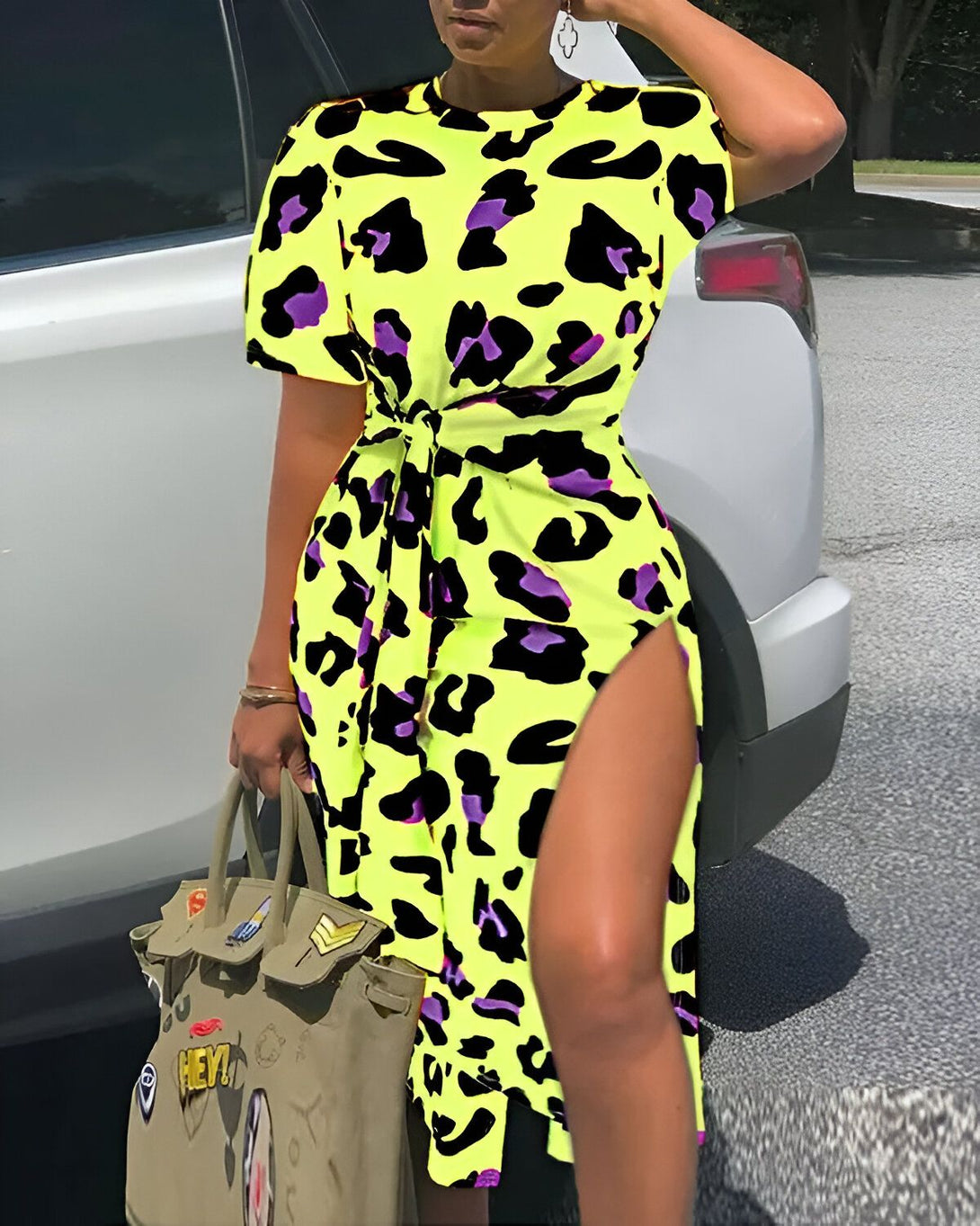 Une femme porte une robe jaune et noire à motifs léopard. La robe est courte et fendue sur le côté. Elle porte également un sac à main vert kaki.
