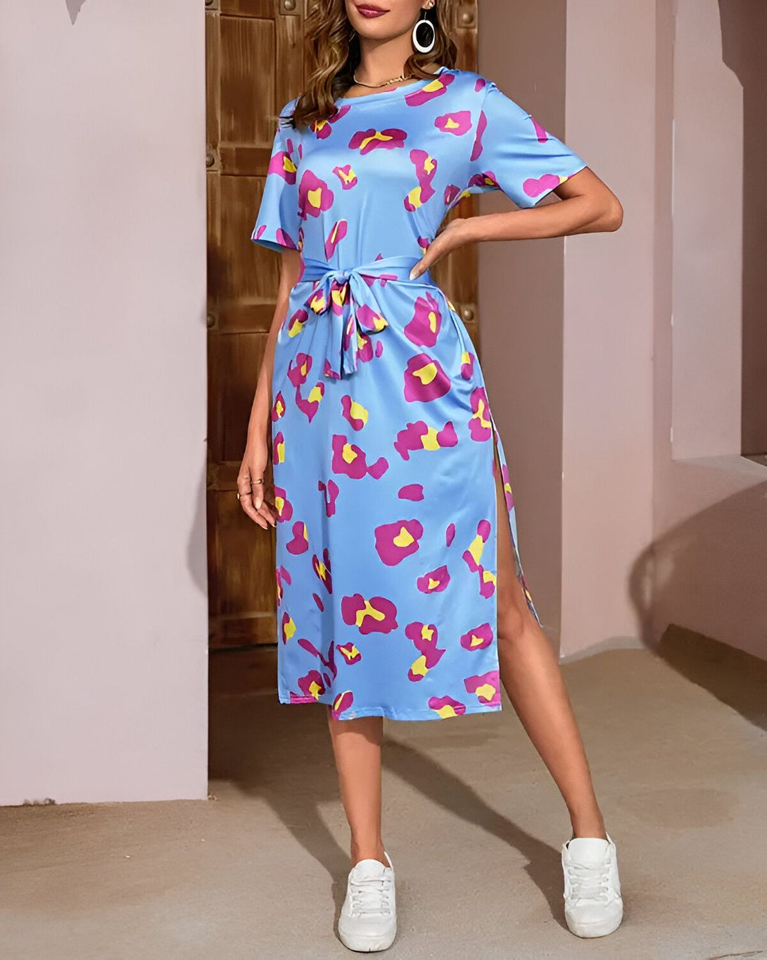 Une femme porte une robe mi-longue portefeuille à imprimé léopard coloré. La robe est ceinturée à la taille et possède une fente sur le côté. Elle est assortie à des baskets blanches.