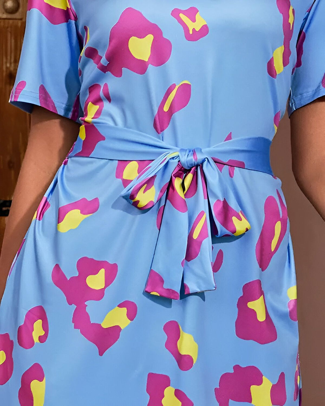 Une robe portefeuille courte à imprimé léopard bleu, rose et jaune. La robe est cintrée à la taille par une ceinture bleue.