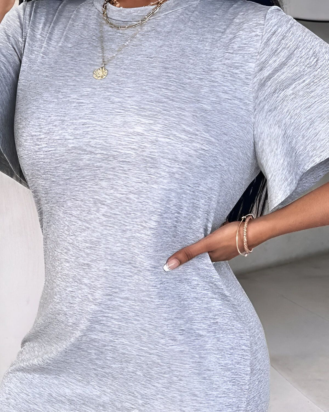 Une femme porte une robe moulante gris chiné à manches courtes. Elle porte également un collier avec un pendentif rond et un bracelet doré au poignet gauche. Ses ongles sont manucurés avec du vernis blanc.