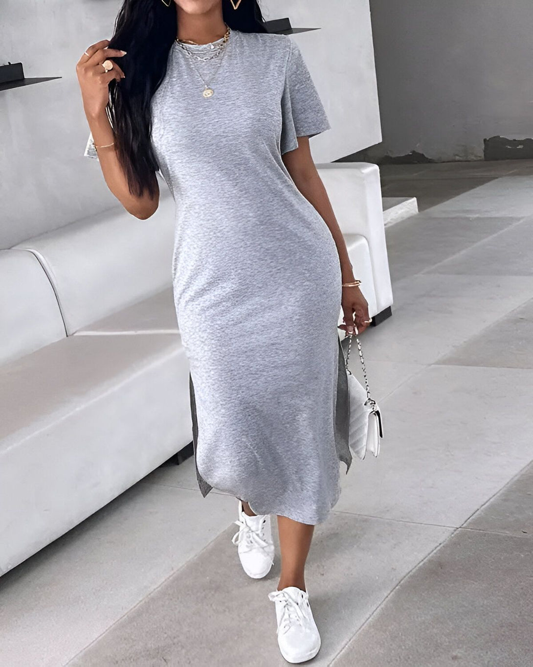 Une femme porte une robe t-shirt grise moulante. La robe est fendue sur les côtés et elle porte des baskets blanches. Elle a les cheveux longs et noirs et porte un collier et des bracelets. Elle est debout dans un salon et il y a un canapé blanc derrière elle.