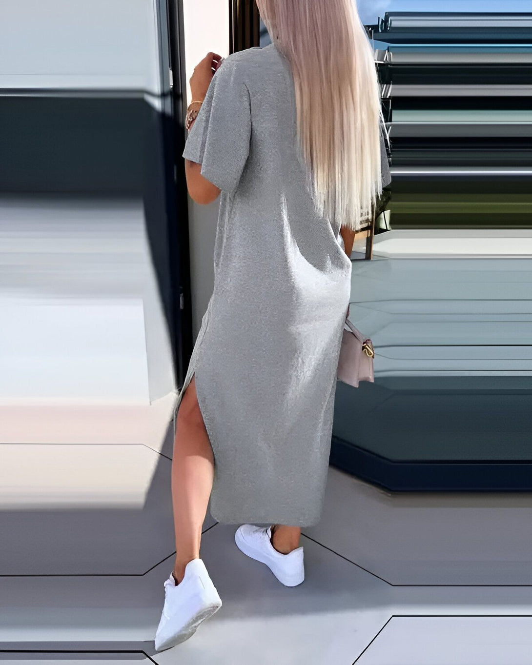 Une femme porte une robe longue grise en jersey. La robe a des manches courtes et une fente sur le côté. Elle porte des baskets blanches et un sac à main rose.