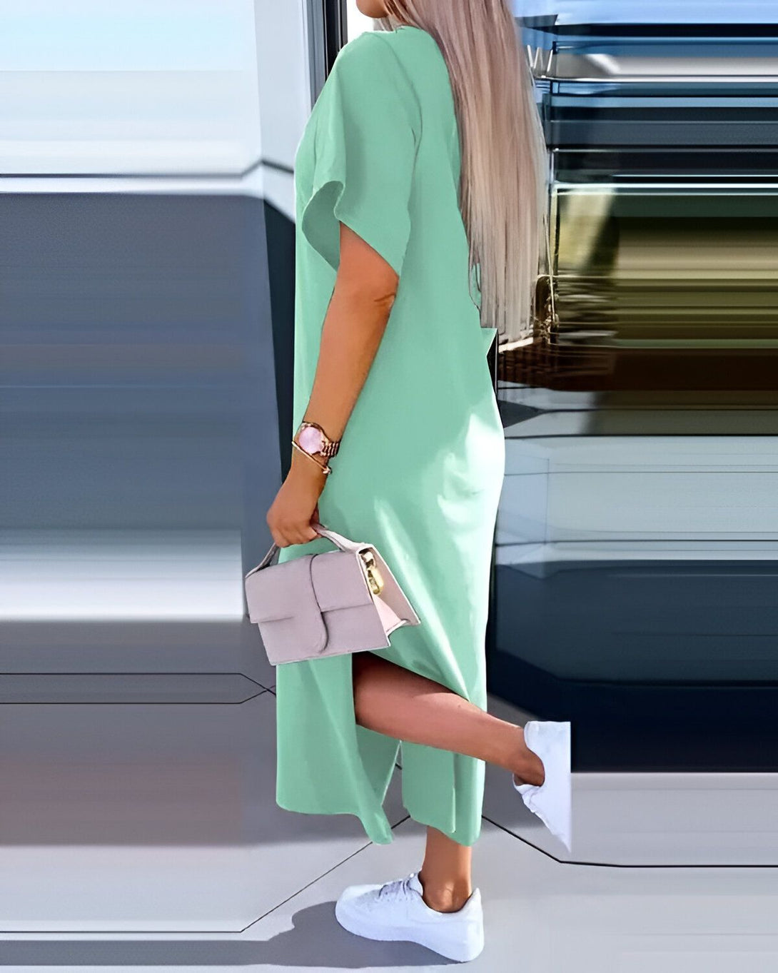 Une femme porte une robe longue vert menthe fendue sur le côté avec des baskets blanches et un sac à main beige.