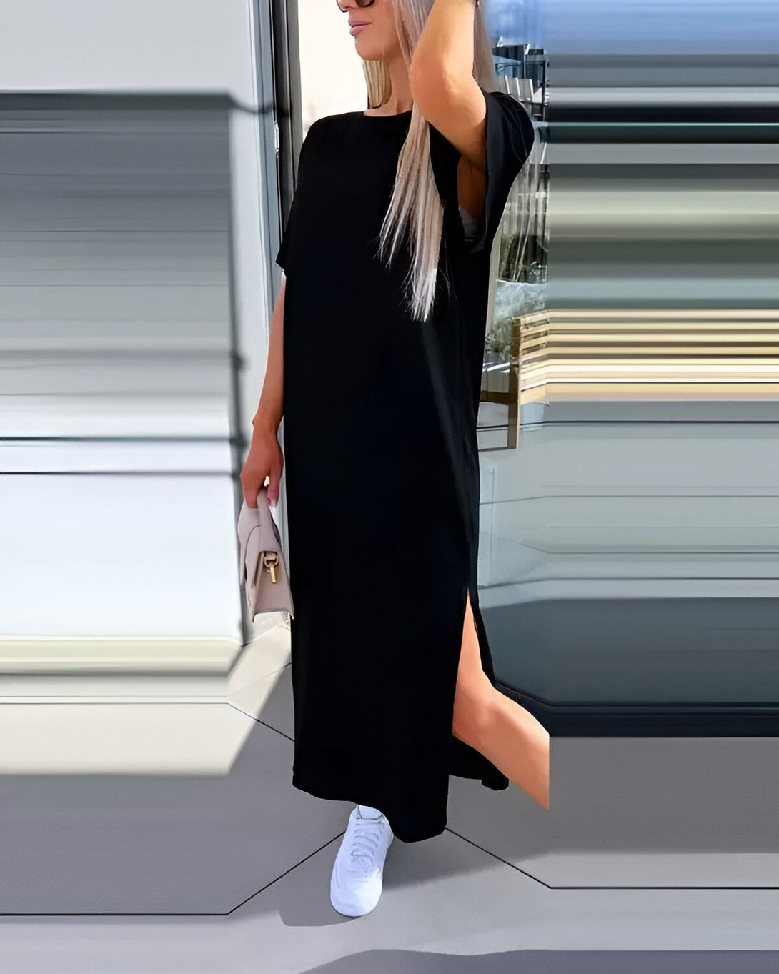 Une femme porte une robe noire longue et ample avec des manches courtes. La robe est fendue sur le côté. Elle porte des baskets blanches et un sac à main beige.