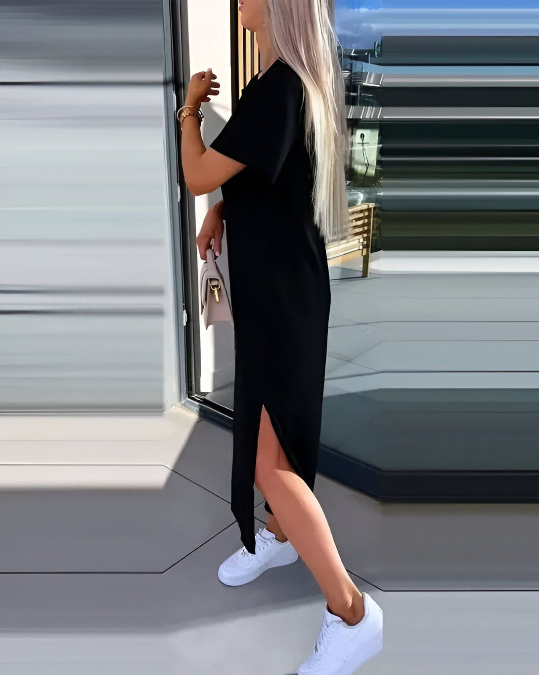 Une femme porte une robe noire fendue sur le côté avec des baskets blanches et un sac à main beige.