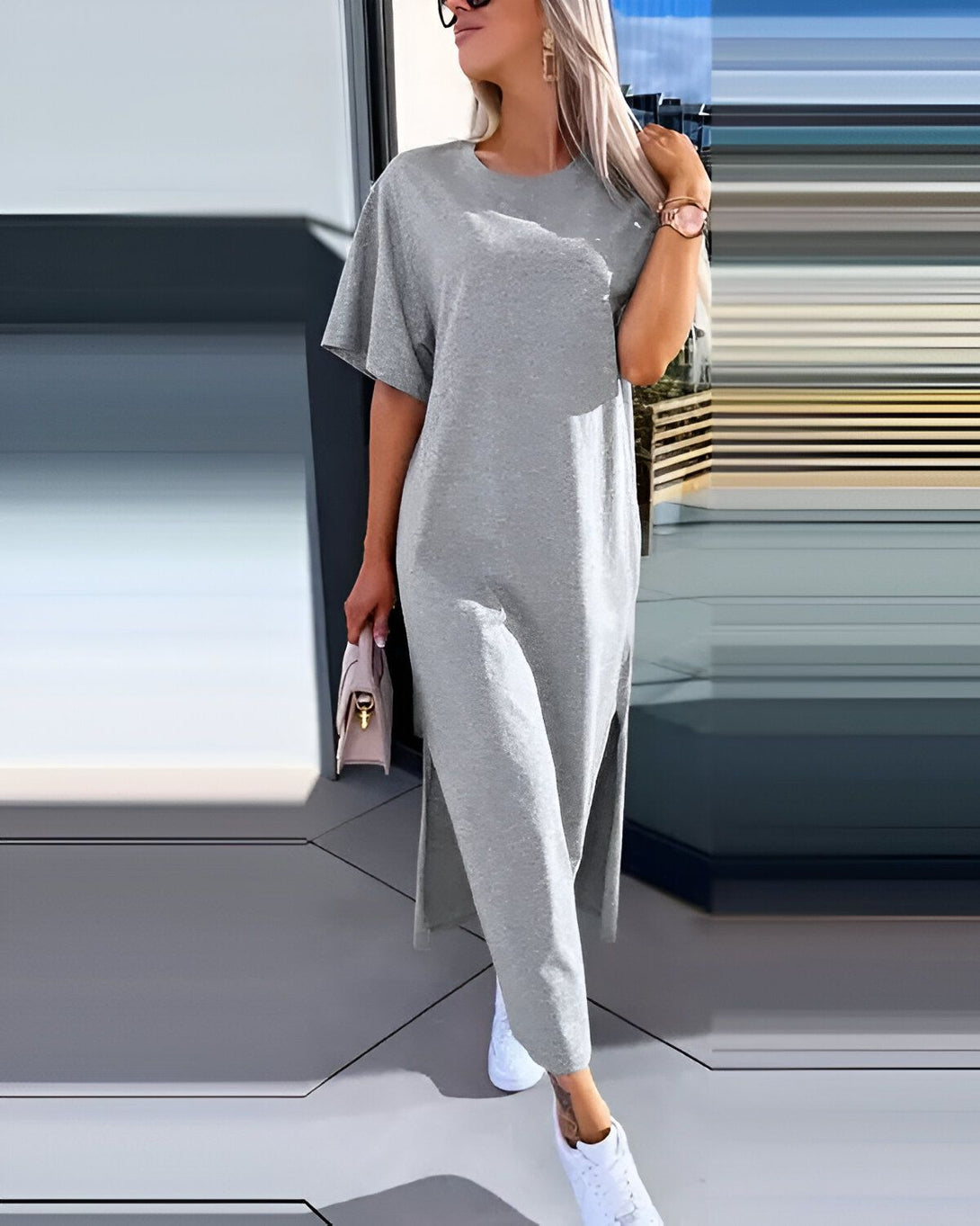 Une femme porte une robe longue en jersey gris clair. La robe a des manches courtes et une fente sur le côté. La femme porte également des baskets blanches et un sac à main beige.