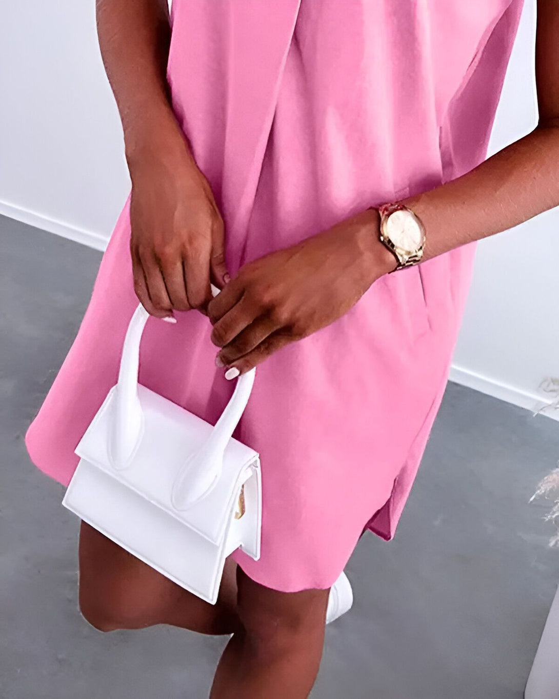 Une femme porte une robe rose et un sac à main blanc. Elle a une montre en or au poignet.