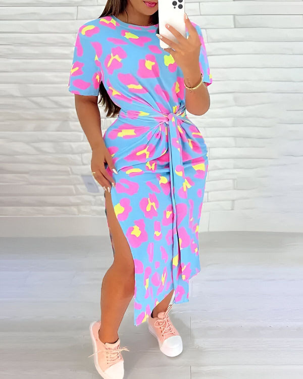 Une femme porte une robe mi-longue moulante à imprimé léopard bleu, rose et jaune. La robe est ceinturée à la taille et possède une fente sur le côté. La femme est debout, de face, et porte des baskets blanches.