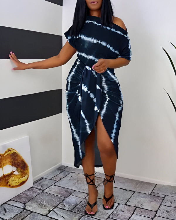 Une femme porte une robe mi-longue portefeuille en wax à nouer sur le côté. La robe est bleu marine et blanche. La femme est noire et a les cheveux longs noirs. Elle porte des sandales noires à talons hauts. Elle est debout dans un coin, la main droite sur le mur et la gauche sur la hanche.