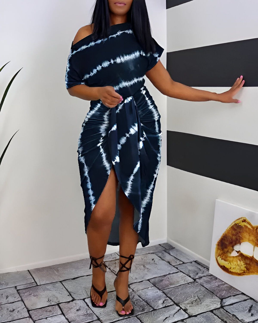 Une femme porte une robe mi-longue tie-dye bleu marine et blanche. La robe a une épaule dénudée et une fente sur le côté. La femme est debout dans un coin, une main sur la hanche et l'autre tenant le mur. Elle porte des sandales à talons hauts noires et des ongles roses.
