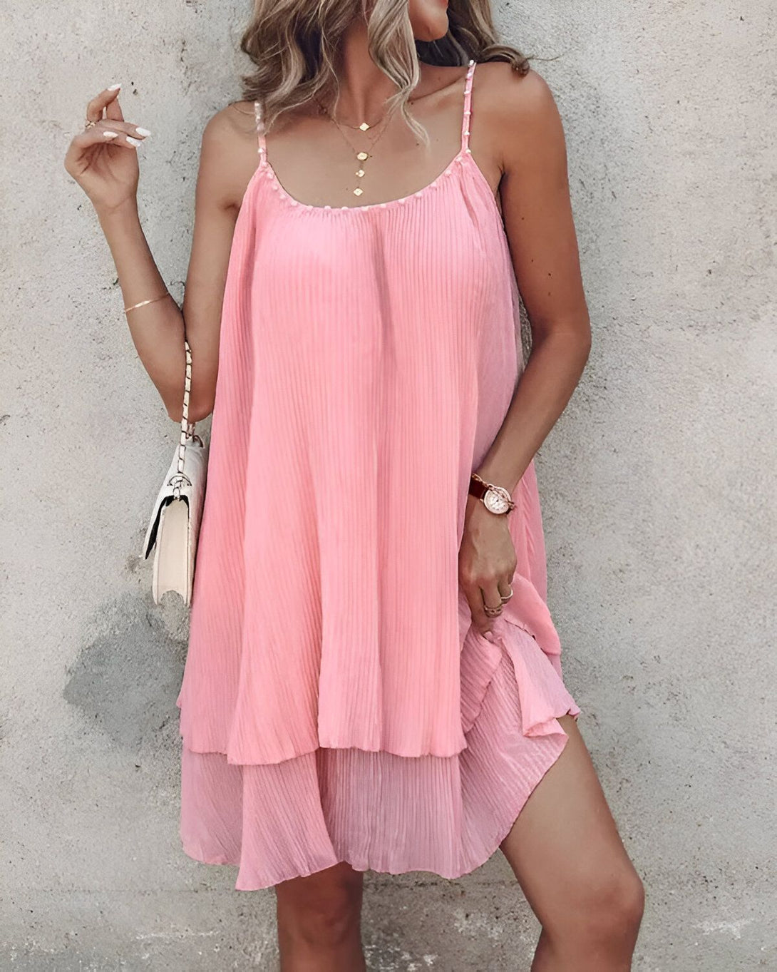 Une jeune femme porte une robe rose à bretelles. La robe est courte et évasée, avec un ourlet à volants. La femme a les cheveux longs et blonds, et elle porte un collier et un bracelet. Elle est debout devant un mur de pierre.