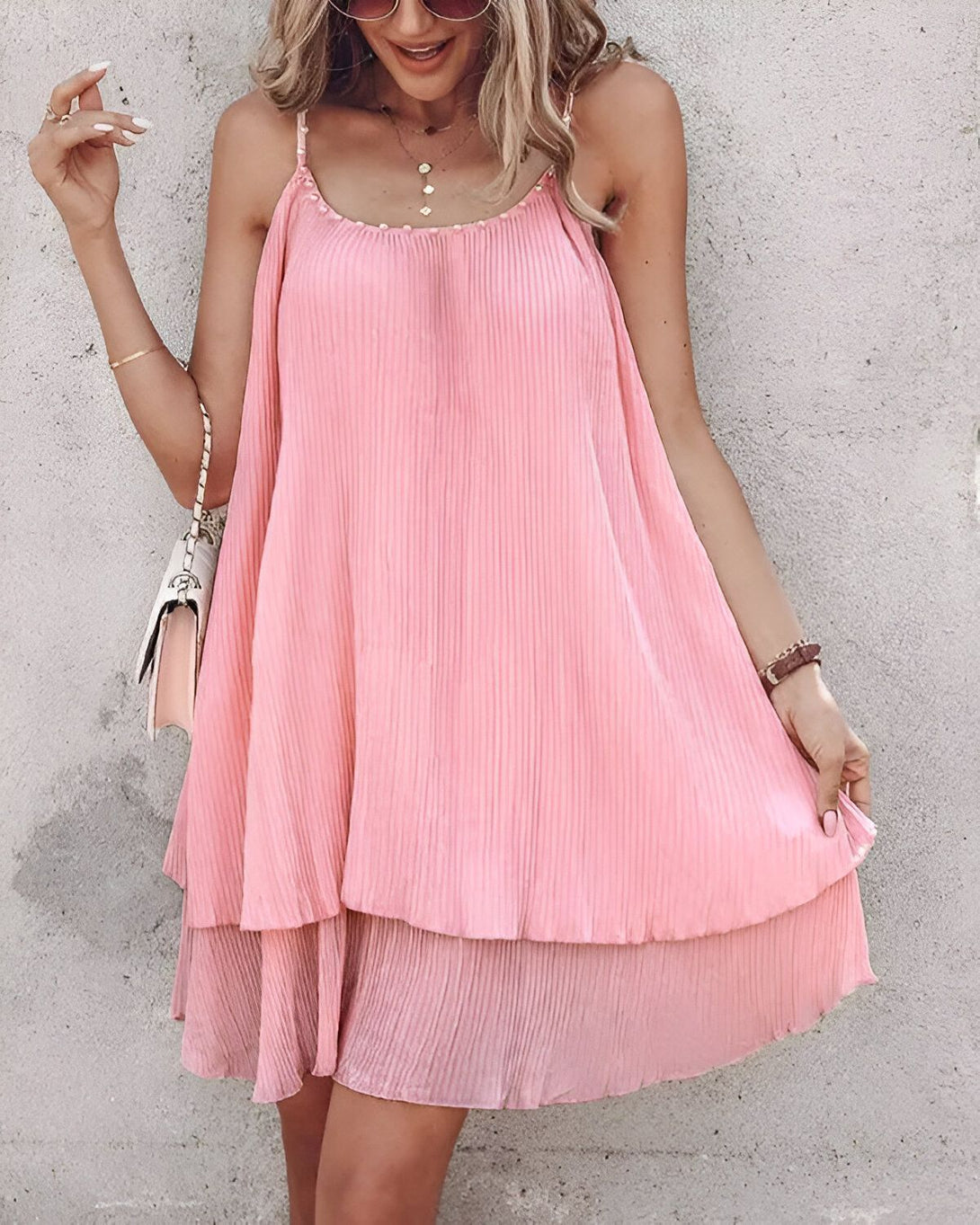 Une jeune femme porte une robe rose plissée. La robe a des bretelles fines et un ourlet évasé. La femme porte également un collier, un bracelet et des lunettes de soleil. Elle a les cheveux longs et blonds et un maquillage léger. Elle se tient debout devant un mur de béton et sourit à la caméra.
