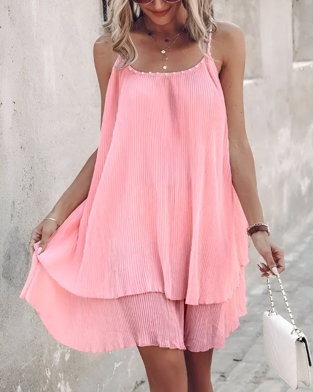 Une femme porte une robe rose plissée et des sandales blanches. Elle a un sac à main blanc à la main.
