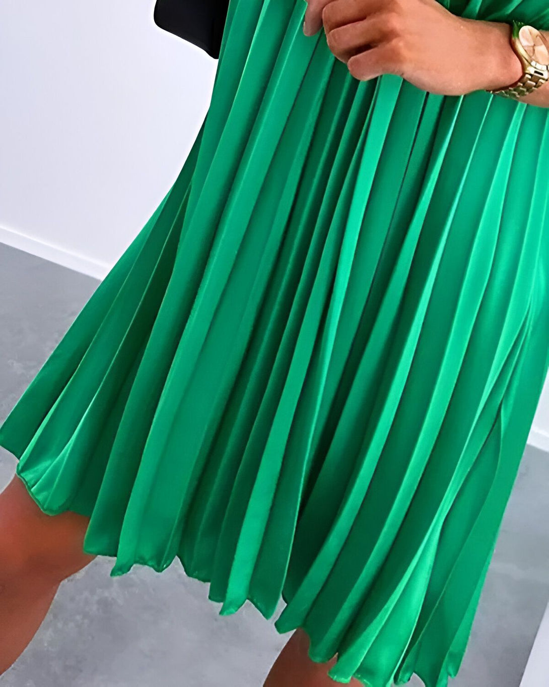 Une femme porte une robe verte plissée. La robe est courte et s'évase légèrement. La femme est également porte une veste noire et des talons hauts noirs.