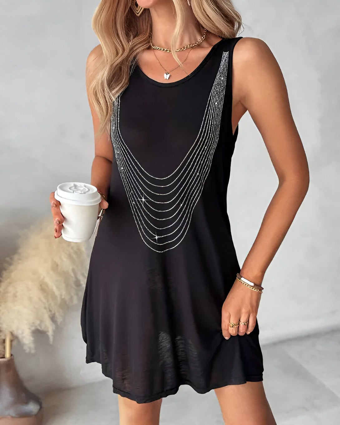 Une femme porte une robe noire sans manches avec un motif de chaînes en strass. Elle a les cheveux blonds lâchés et porte un collier avec un pendentif en forme de papillon. Elle tient une tasse blanche à la main.