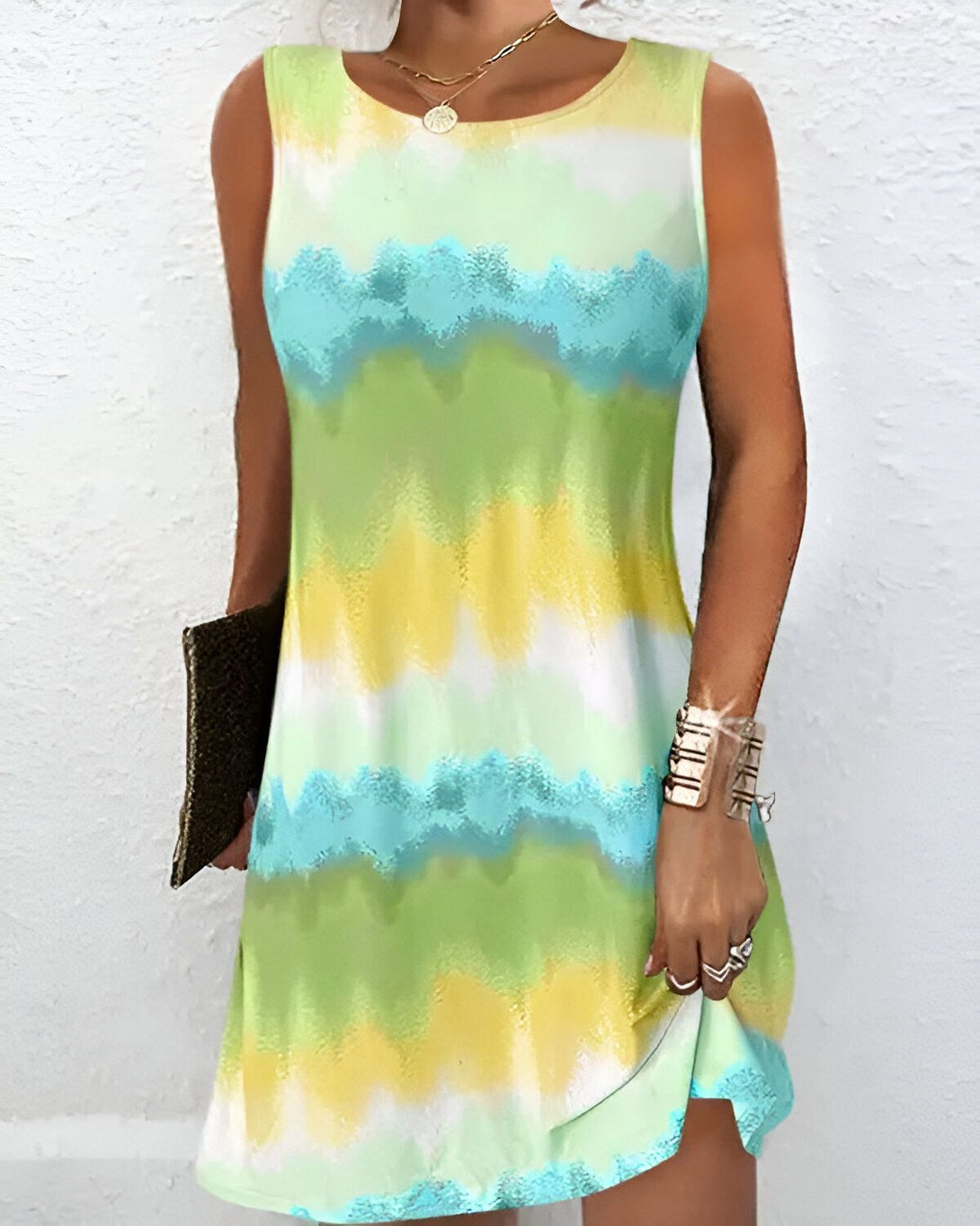Une jeune femme porte une robe verte, jaune et bleue sans manches. La robe est courte et évasée. La femme porte également un collier et des bracelets. Elle a un sac à main marron à la main.