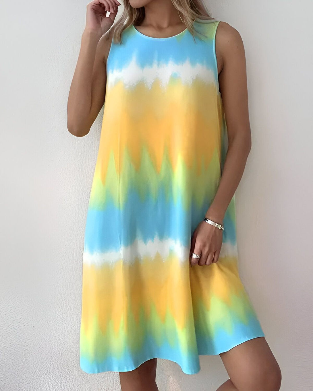 Une femme porte une robe jaune, bleue et verte tie-dye. La robe est sans manches et a une encolure ronde. La femme est debout et a la main sur la hanche. Elle porte également un bracelet en argent au poignet gauche.
