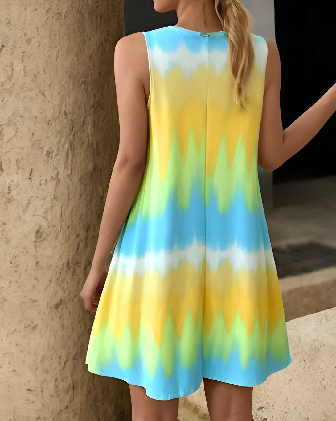 Une femme porte une robe jaune, verte et bleue sans manches. La robe est courte et évasée. La femme est blonde et a les cheveux longs. Elle est vue de dos.
