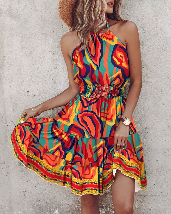 Une femme porte une robe colorée à motifs cachemire. La robe est courte et évasée, avec un haut dos nu et une taille froncée. La femme est debout devant un mur de béton et porte un chapeau de paille.