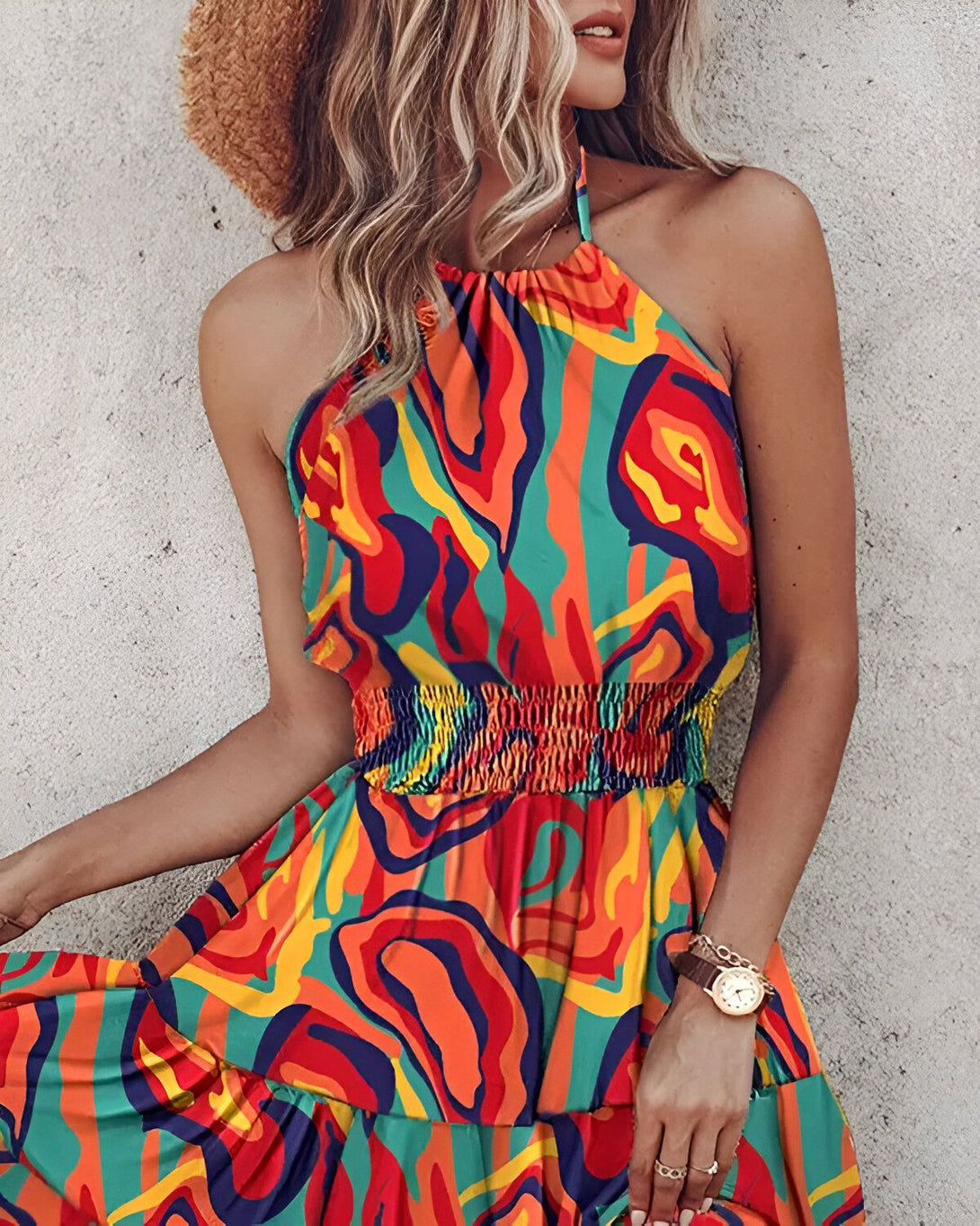 Une jeune femme porte une robe colorée à motifs. La robe est courte et évasée, avec un haut halter et une taille froncée. La femme porte également un chapeau de paille et des sandales.