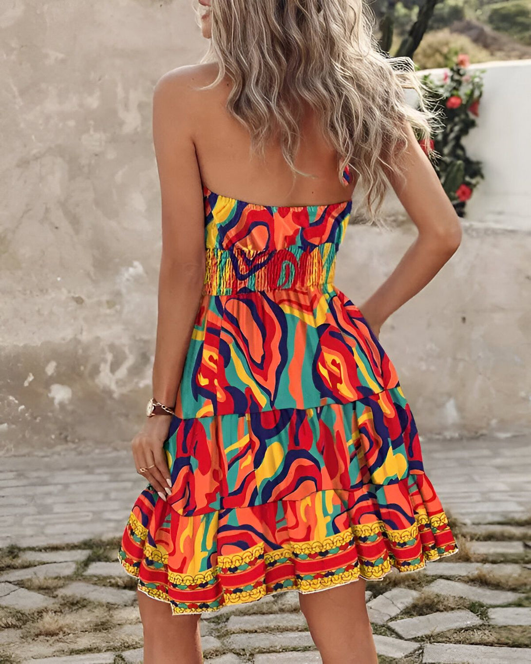 Une femme porte une robe bustier colorée à motifs. La robe est courte et évasée, avec un volant dans le bas. La femme est blonde et a les cheveux longs. Elle est vue de dos.
