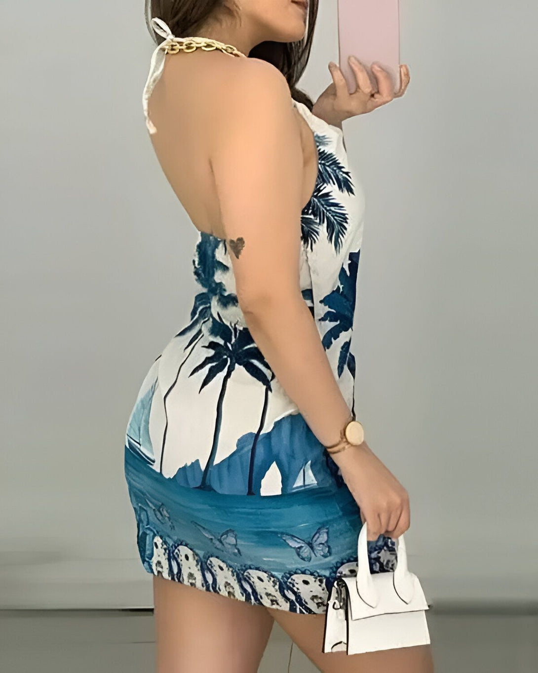 Une femme porte une robe d'été blanche et bleue. La robe est courte et moulante. Elle a un imprimé de feuilles de palmier et de fleurs. La femme est vue de dos. Elle a les cheveux longs et bruns. Elle porte un collier et un bracelet. Elle tient un sac à main blanc.