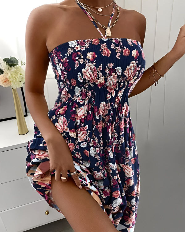 Une jeune femme porte une robe bustier à fleurs. La robe est courte et évasée, elle est bleu marine avec des fleurs roses et rouges. La femme est debout, elle a les cheveux longs et bruns et elle porte un collier.