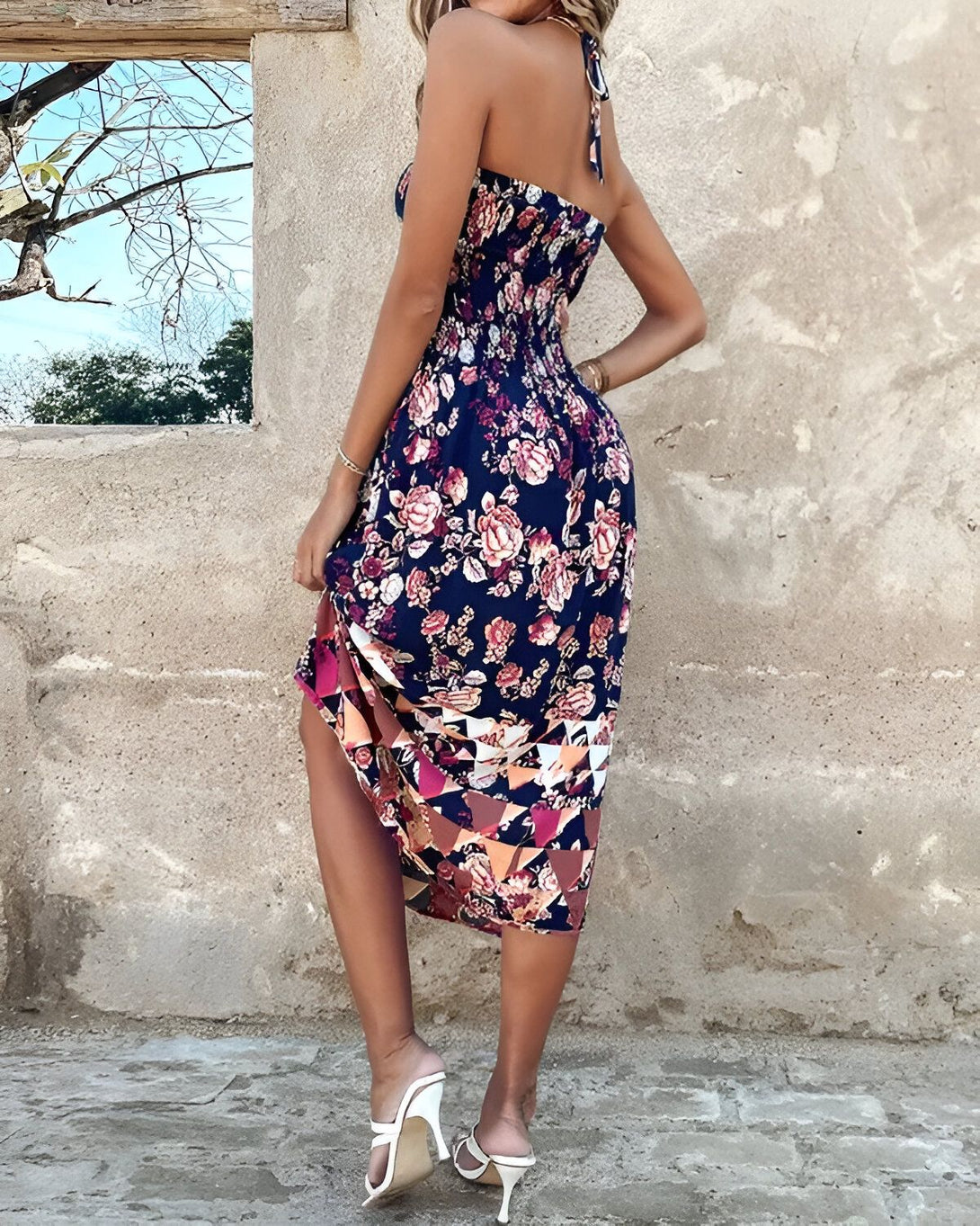 Une femme porte une robe longue bleu marine à motifs floraux. La robe est fendue sur le côté et l'on peut voir ses jambes. Elle porte des sandales à talons hauts blanches.
