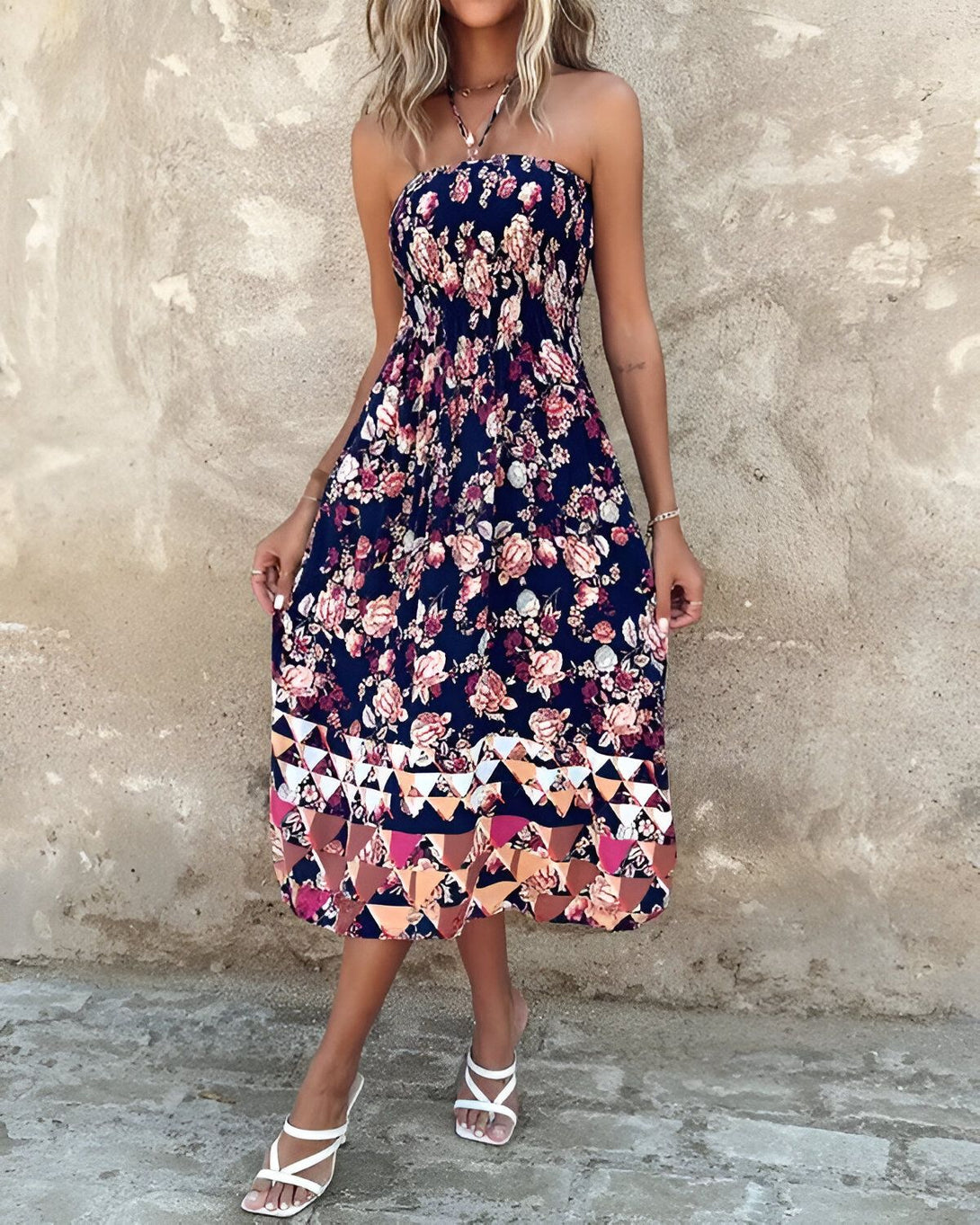 Une femme porte une robe mi-longue à fines bretelles. La robe est bleu marine à motifs fleuris roses et rouges. Le bas de la robe est orné de motifs géométriques multicolores. La femme est blonde et porte des sandales blanches. Elle se tient debout devant un mur de pierre.