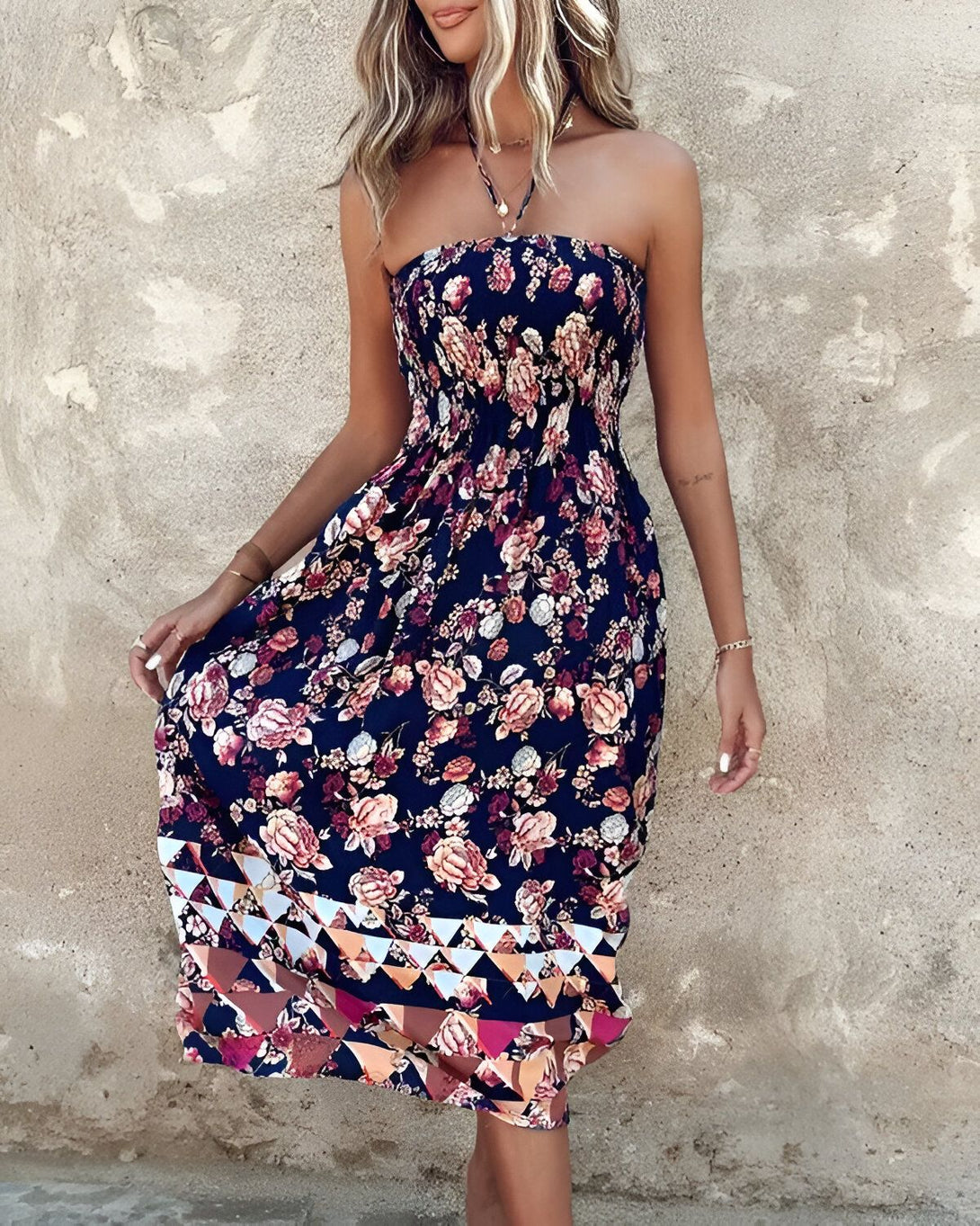 Une jeune femme porte une robe longue et colorée sans bretelles. La robe est à motifs floraux et géométriques. La femme est blonde et souriante. Elle porte un collier et des bracelets. Elle est pieds nus.