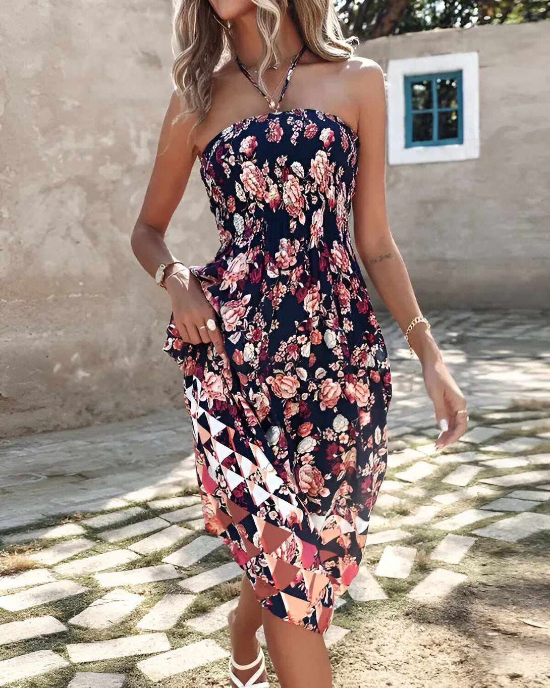 Une femme porte une robe longue et fleurie avec un décolleté en V et des bretelles fines. La robe est bleu marine avec des fleurs roses et rouges. La femme est blonde et porte des sandales blanches. Elle se trouve dans un jardin et sourit.