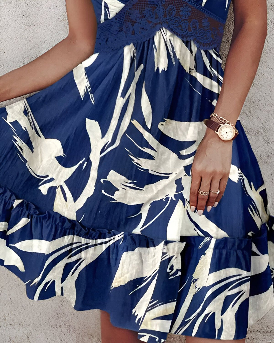 Une femme porte une robe bleue à motifs blancs. La robe est courte et évasée, avec des volants dans le bas. La femme est également porte une montre marron et une bague en or à la main droite.