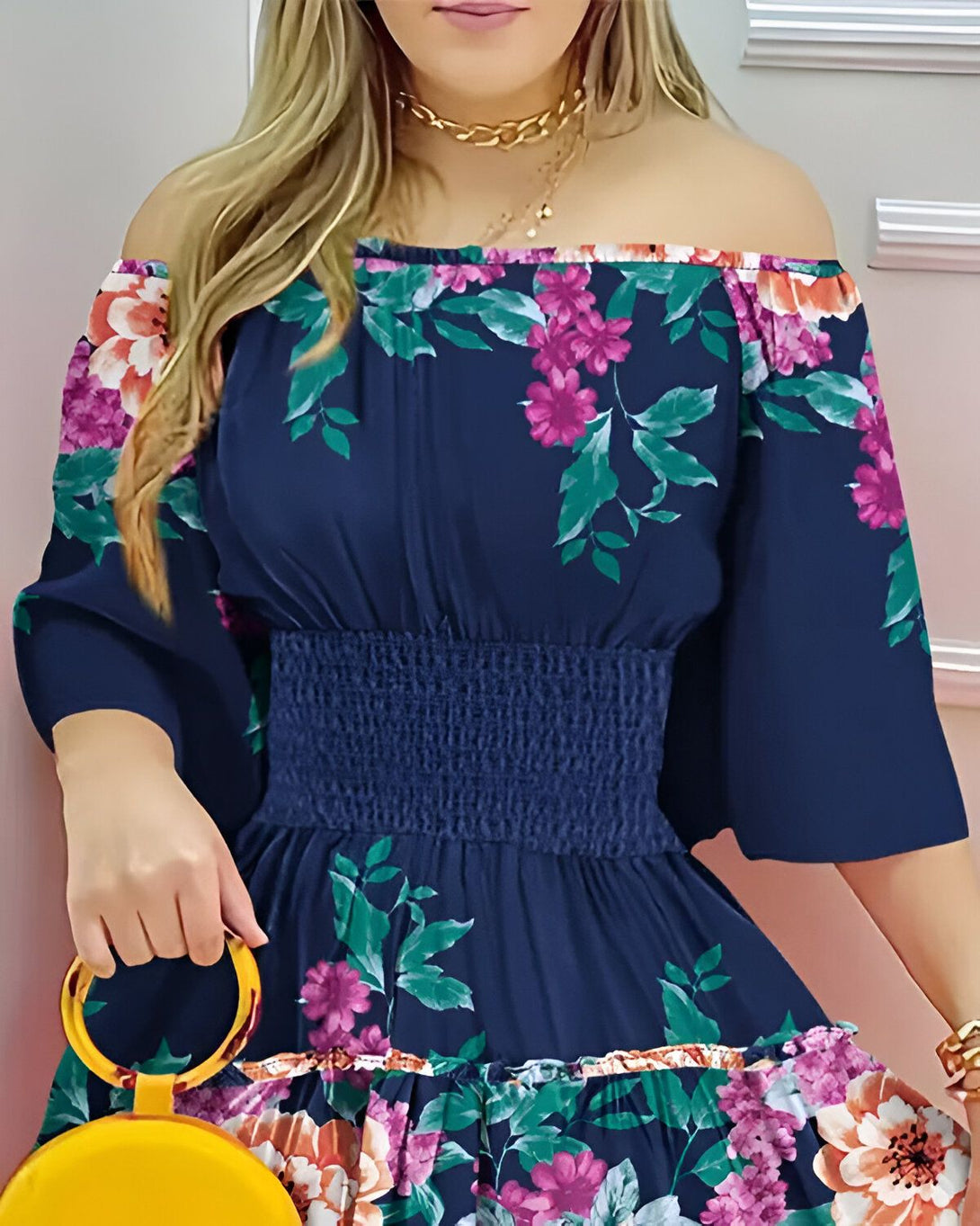Une jeune femme porte une robe bleu marine à fleurs roses et violettes. La robe est à épaules dénudées et cintrée à la taille. La femme porte également un collier doré et un sac à main jaune.
