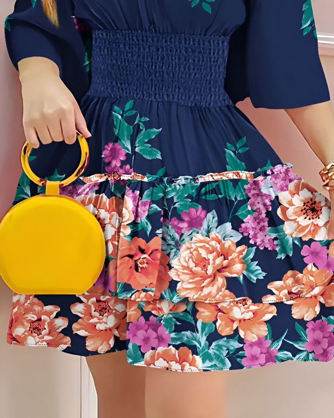 Une femme porte une robe bleu marine à fleurs et un sac à main jaune. La robe est courte et évasée, avec un élastique à la taille. Le sac à main est rond et a une anse jaune. La femme est debout et tient le sac à main dans sa main droite.