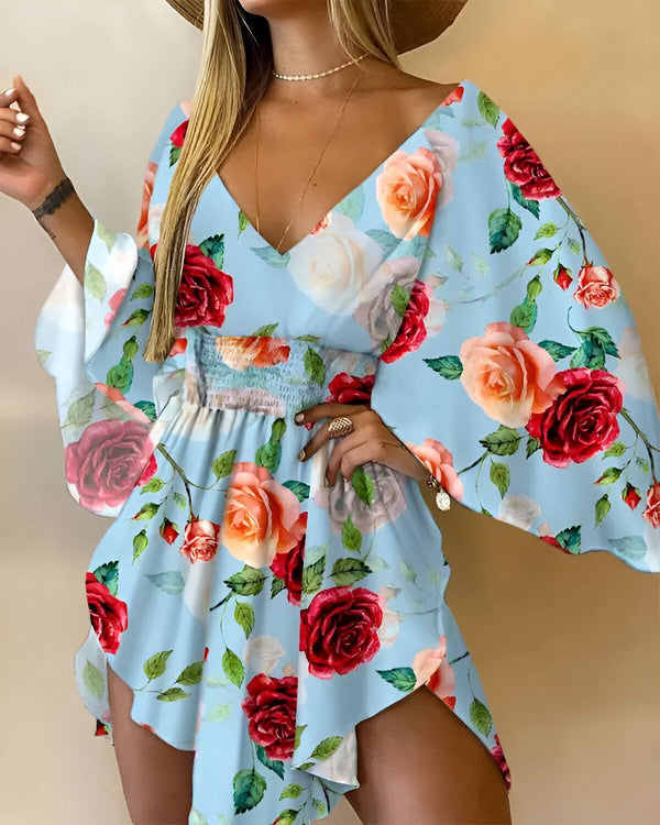 Une femme porte une robe bleu clair à motifs floraux rouges et roses. La robe est courte et évasée, avec des manches bouffantes. La femme porte également un chapeau de paille marron et des sandales marron.
