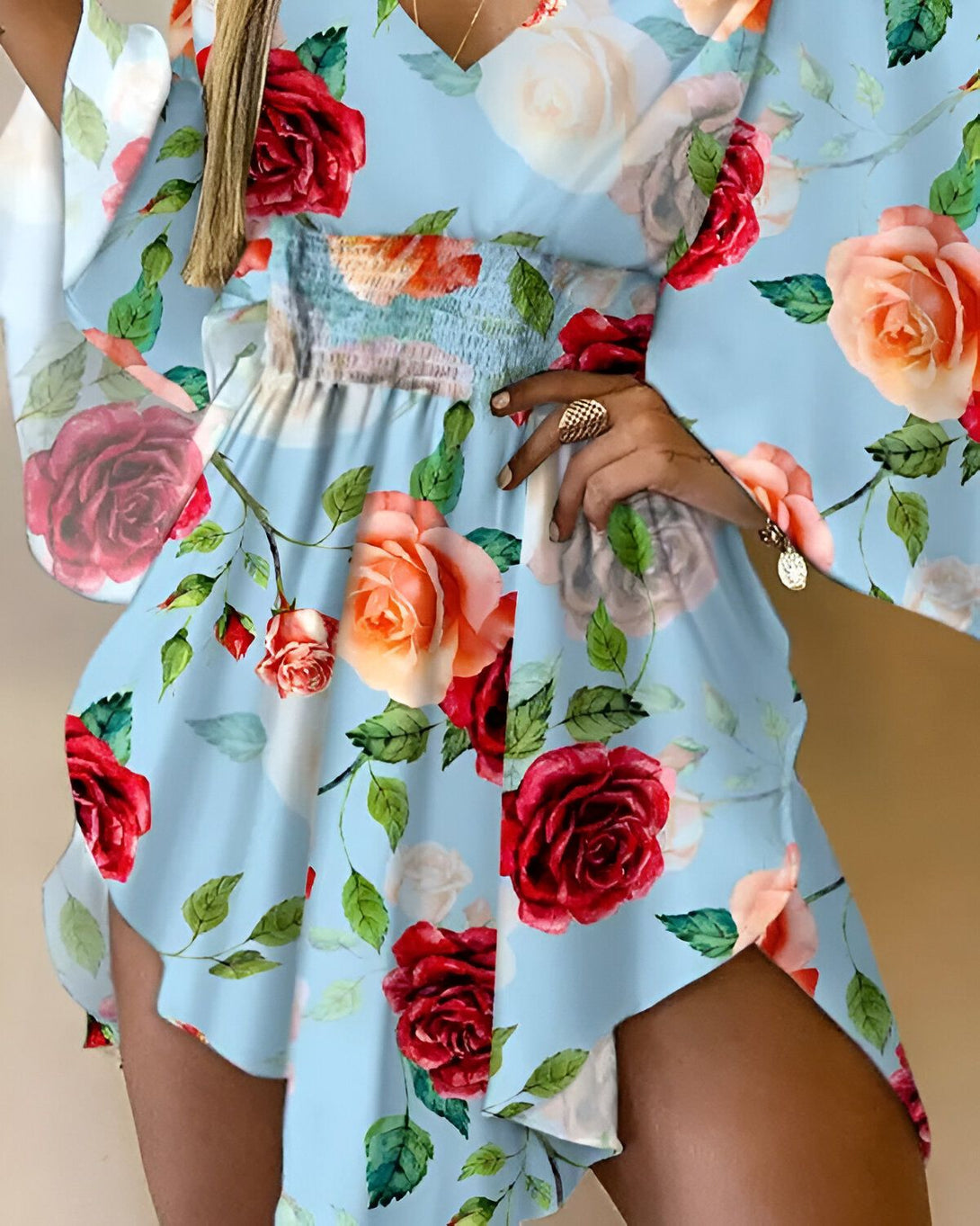 Une femme porte une robe portefeuille courte à imprimé floral. La robe est bleu clair avec des roses rouges et des feuilles vertes. La femme est debout, les mains sur les hanches.