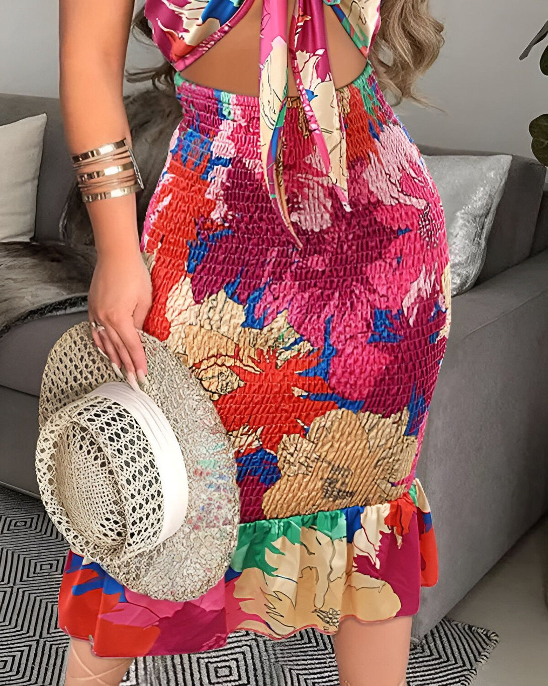 Une femme porte une robe longue rose et blanche à motifs floraux. Elle est debout dans un salon et tient un chapeau de paille à la main.