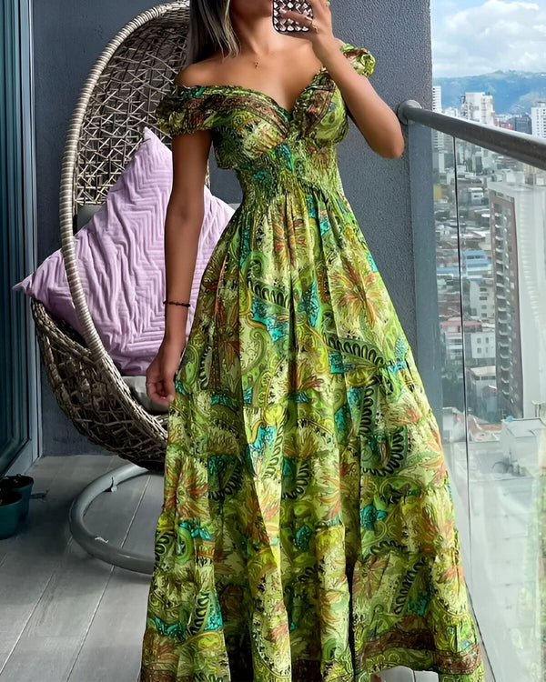 Une femme est debout sur un balcon et porte une robe longue verte à motifs. La robe est décolletée et a des manches courtes. La femme est pieds nus et porte un collier. Elle a les cheveux longs et noirs. En arrière-plan, on peut voir une ville.