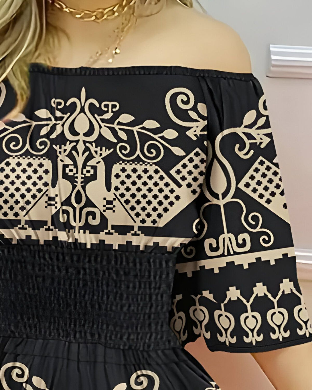 Une femme porte une robe noire à motifs blancs. La robe est sans manches et a une encolure Bardot. La femme est blonde et porte un collier doré.