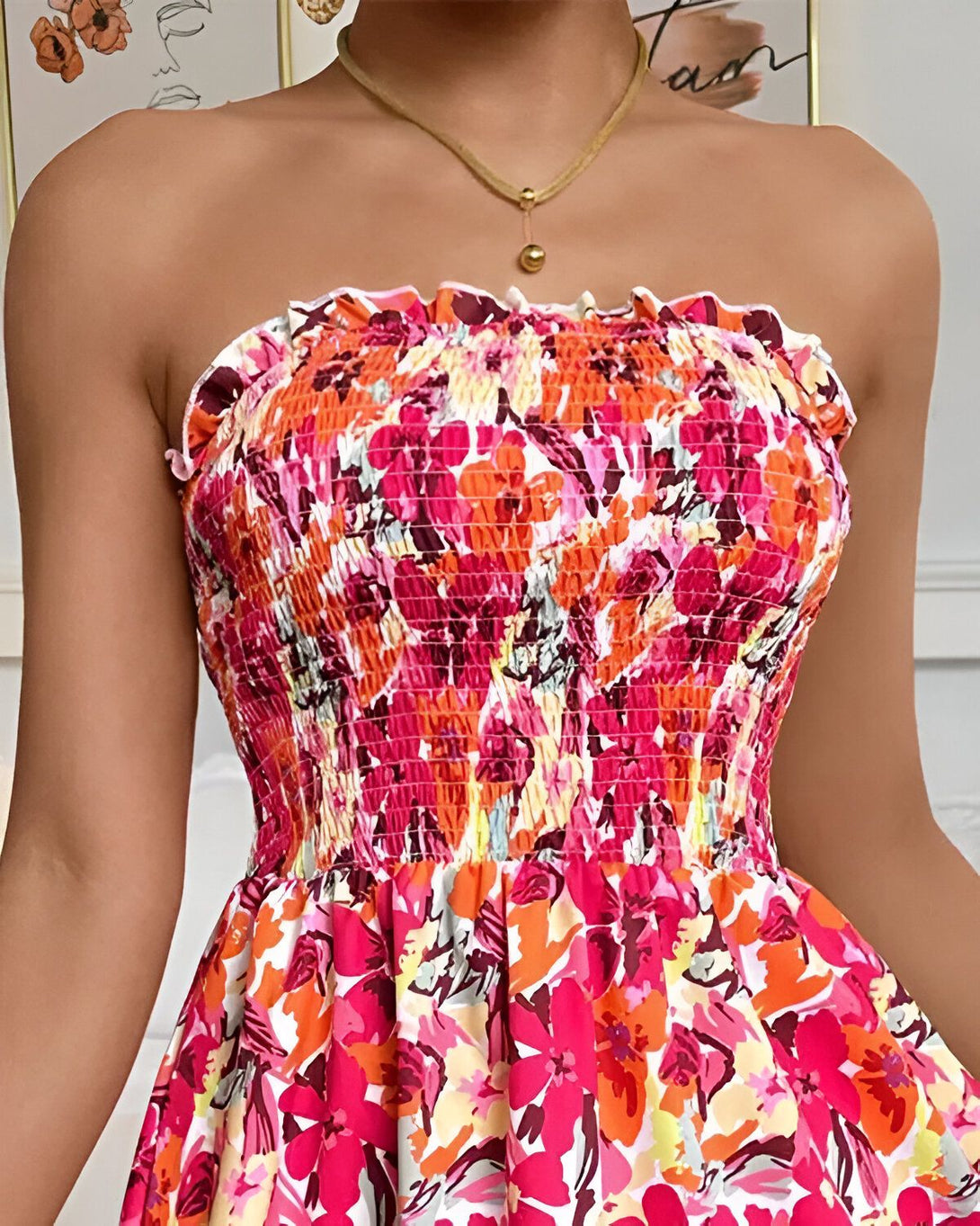 Une jeune femme porte une robe bustier à fleurs. La robe est rose, orange et blanche. La femme est souriante et porte un collier doré.